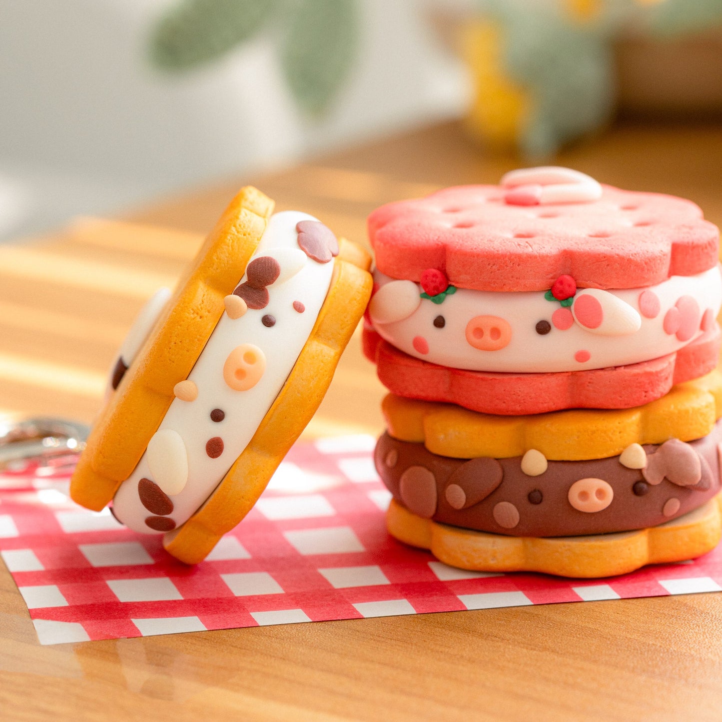 Three cute, cozy cow-themed sandwich cookies with a keychain ring, displayed on a red and white checkered napkin on a wooden table with a soft-focus background.