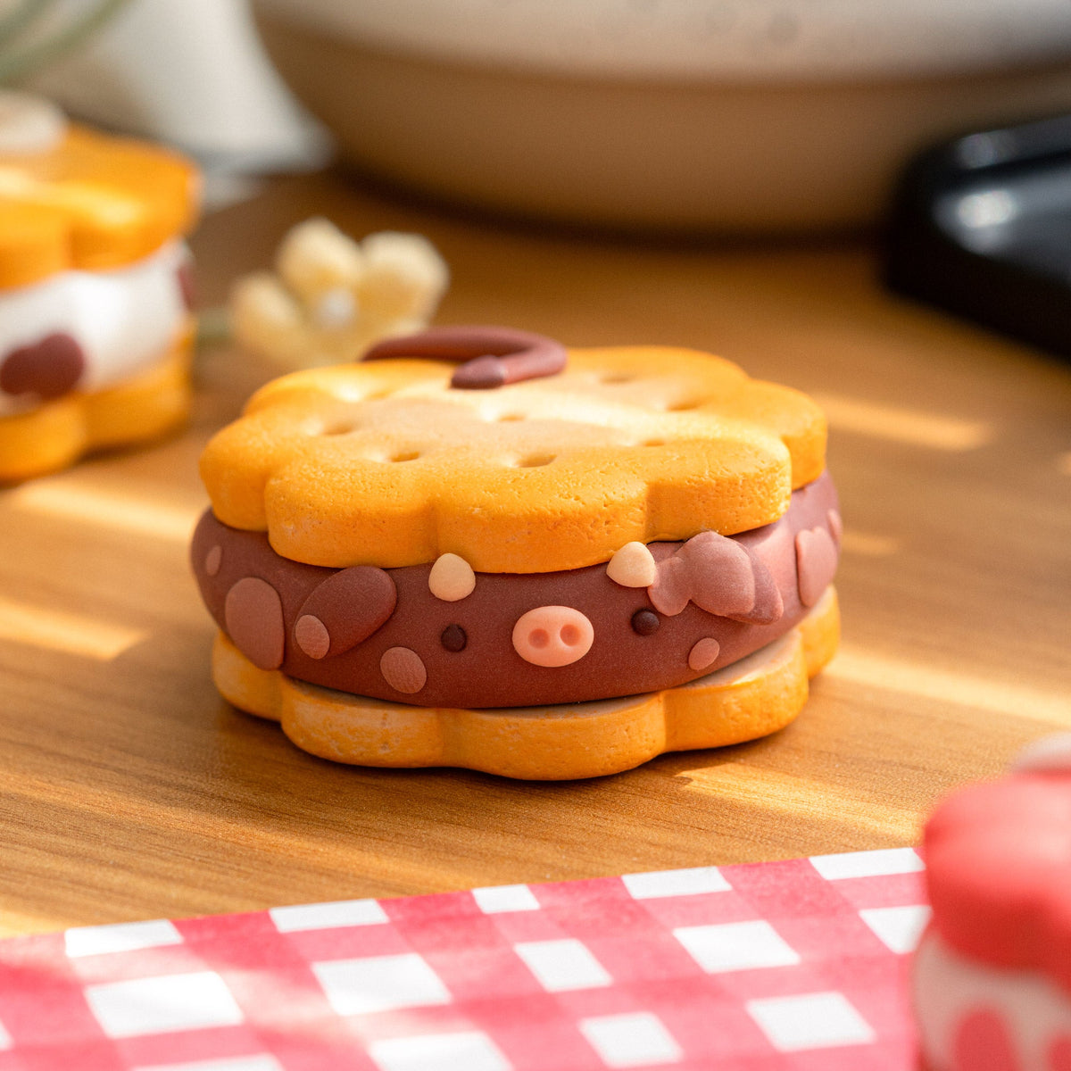 A close-up of a small cow-shaped sandwich cookie with a chocolate-like brown filling decorated with pink and beige spots, set on a wooden surface with a red-and-white checkered cloth in the foreground.