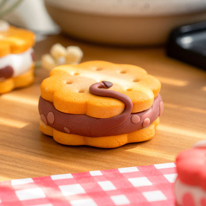 A small decorative sandwich cookie or biscuit-shaped item with a wavy yellow-orange top and bottom and a dark brown cow-shaped center, styled on a wooden surface with a red-and-white checkered cloth and a blurred background.