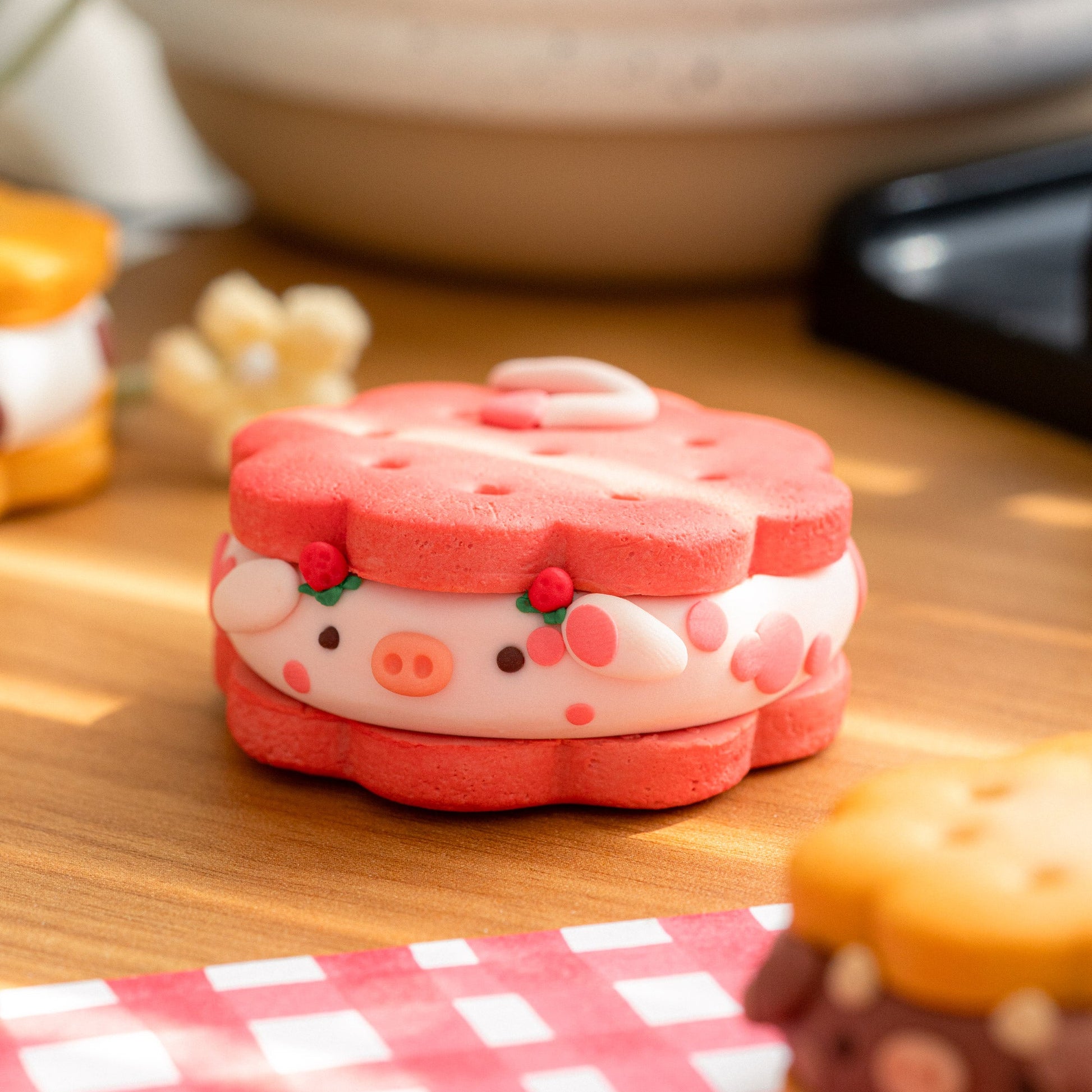 A cute cow-shaped sandwich cookie or clay decoration sits on a wooden table, styled with a red-and-white checkered napkin and other similar treats in the foreground and background.