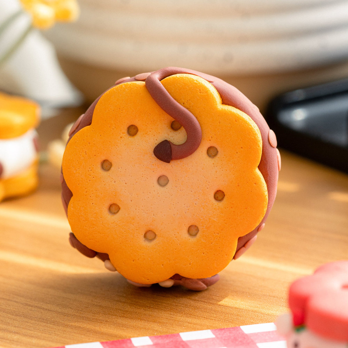 A polymer clay chocolate sandwich cookie with orange cookie layers, a dark brown center, and brown icing details, displayed on a wooden surface with a red-and-white checkered cloth and blurred background props.