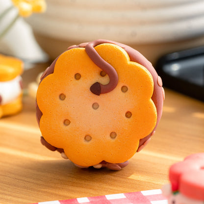 A polymer clay chocolate sandwich cookie with orange cookie layers, a dark brown center, and brown icing details, displayed on a wooden surface with a red-and-white checkered cloth and blurred background props.