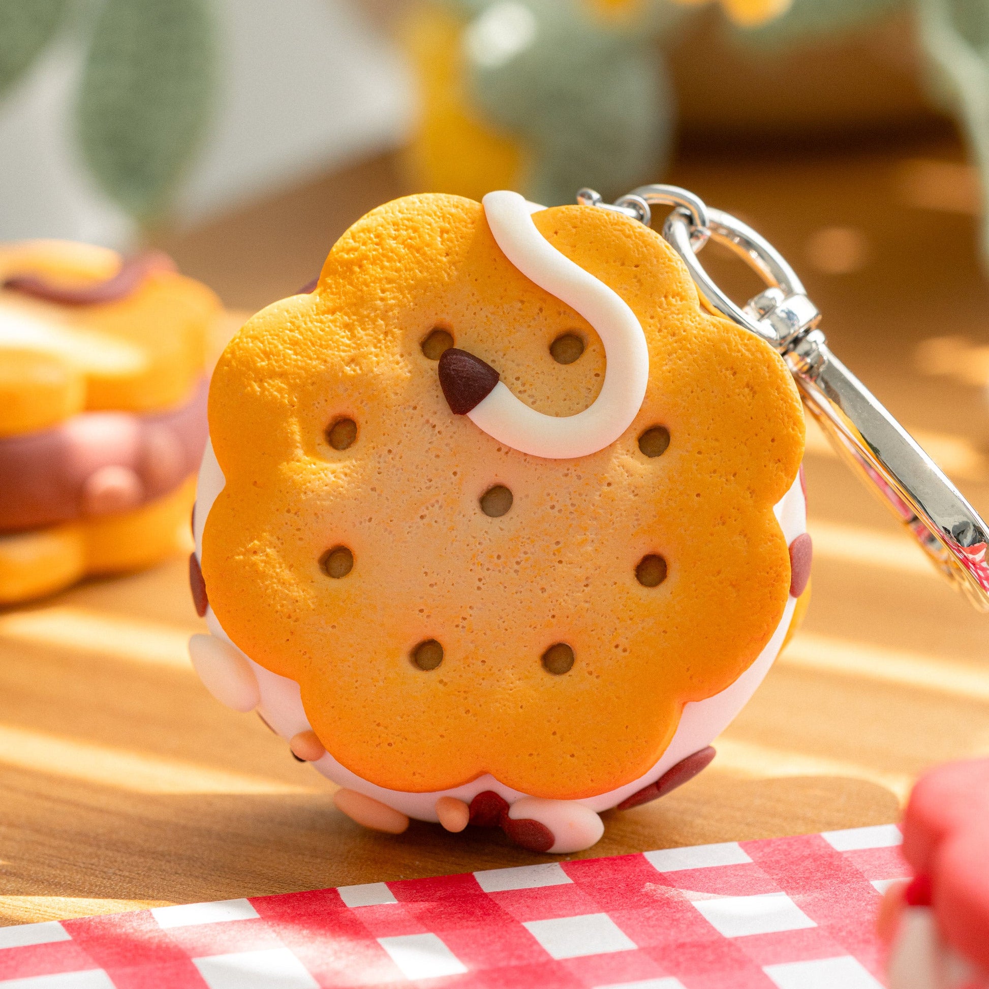 A small cookie-shaped keychain charm with a silver metal key ring, photographed on a wooden surface with a red-and-white checkered cloth in the foreground and a softly blurred background.