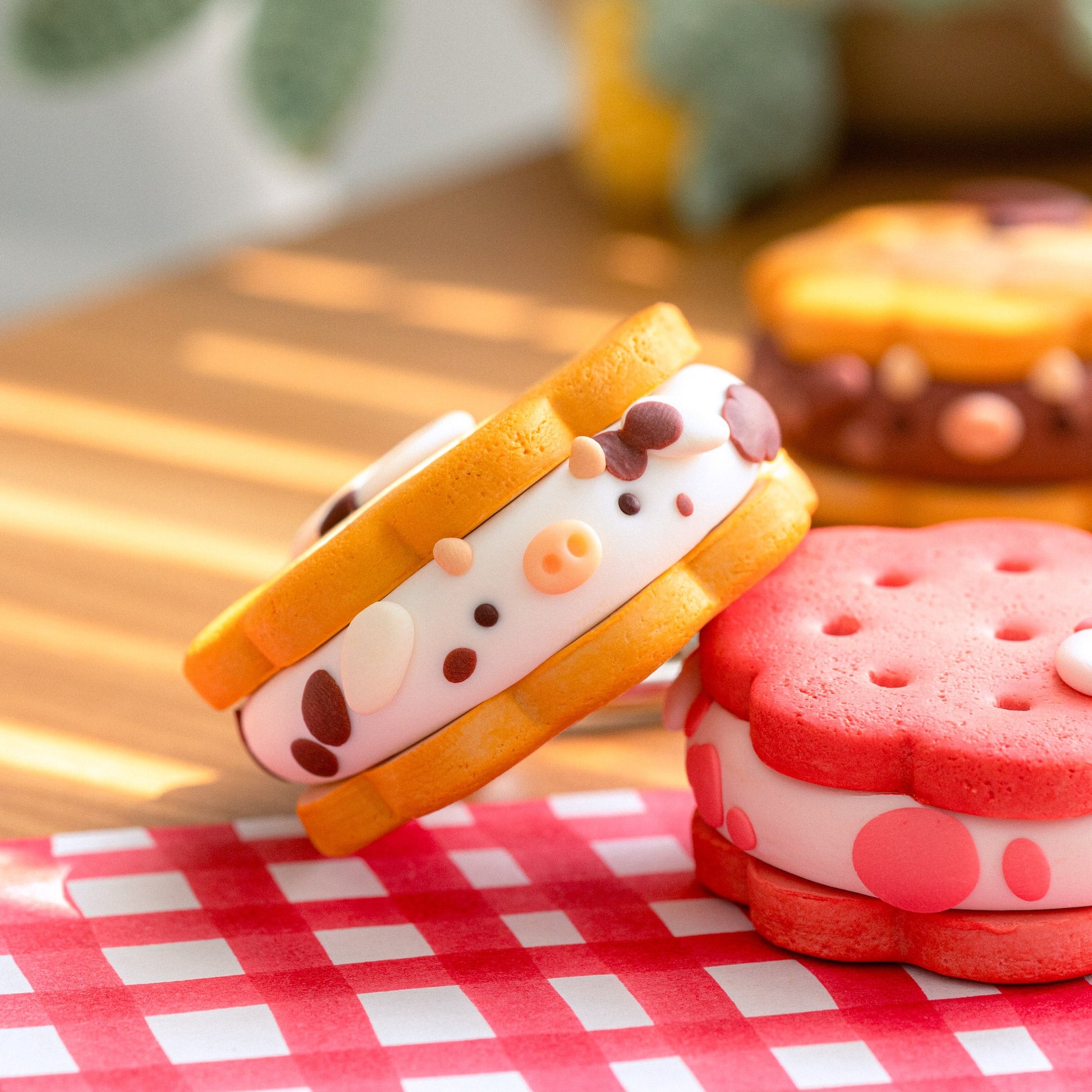 Two cute, cartoon-style sandwich cookies styled like cow faces, one standing upright and the other leaning on a red-and-white checkered napkin on a wooden table.