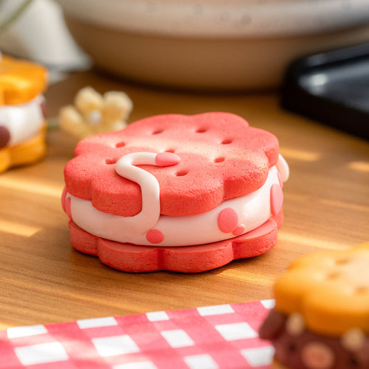 A small pink and white sandwich cookie charm or figurine sits on a wooden surface with a red-and-white checkered cloth in the foreground and other blurred toy-like items in the background.