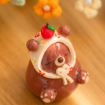 A handmade brown bear figurine and desk friend wearing a cream-colored hood with red and yellow flower accents, sitting on a warm wooden surface with soft blurred flowers in the background.