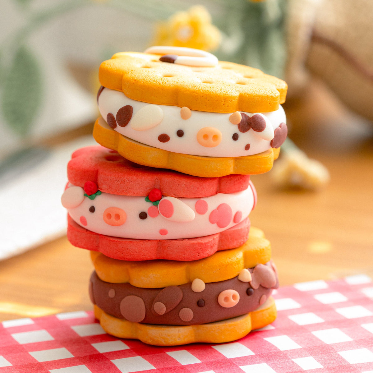Three cute, small clay sandwich cookies styled like cows, stacked on a red and white checkered napkin on a wooden table with a blurred background.